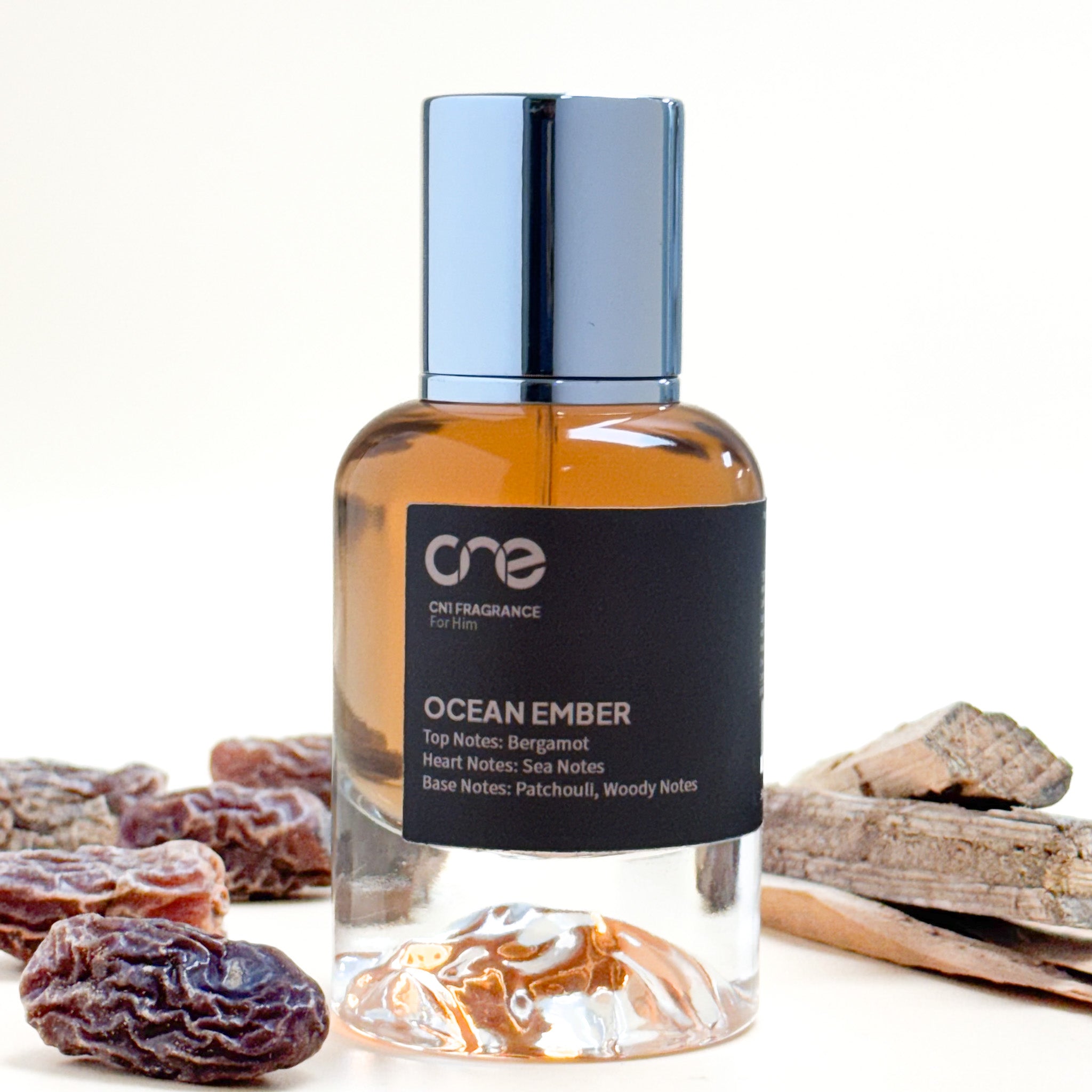 Perfume bottle labeled 'One' with 'Ocean Ember' description, surrounded by dried fruits and driftwood on a white background.